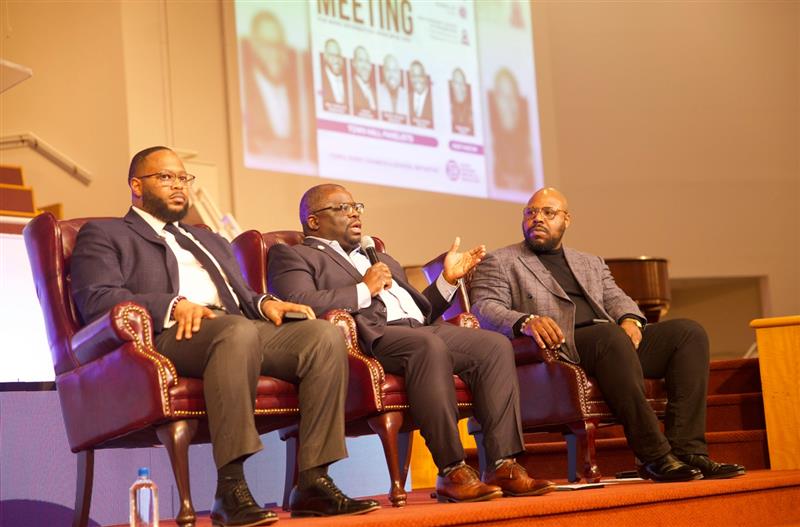 three men seated on a stage participate in a panel discussion, one speaking into a microphone while the others listen, with a presentation screen visible behind them.