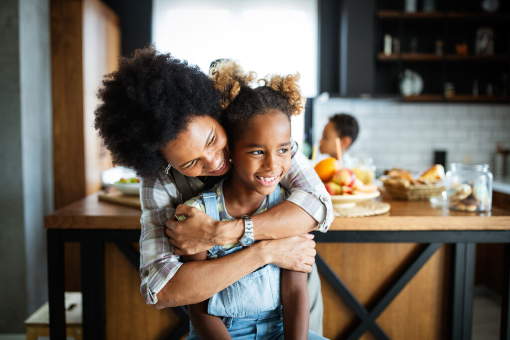 a parent hugs a smiling child in a warm kitchen setting, with food and family activity in the background